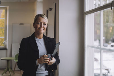 Smiling female professor holding laptop standing by window in innovation lab