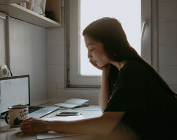 Side view of woman using laptop abd writing notes at home