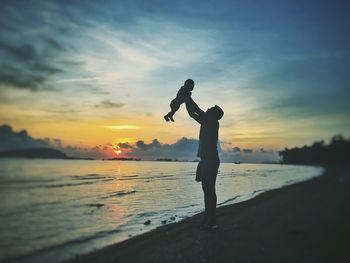 Silhouette father lifting baby while standing at shore during sunset
