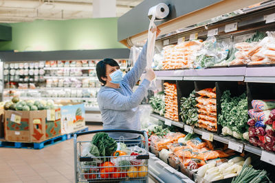 Middle age woman in face mask tearing off plastic transparent disposable bag in grocery store shop 