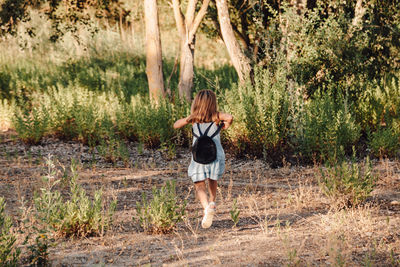 Rear view of girl with backpack running on field against plants in forest