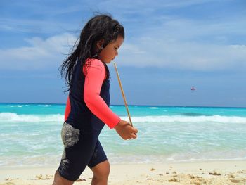 Side view of girl holding stick while walking at beach against sky