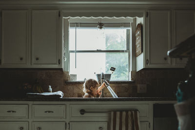 Playful girl playing with faucet while sitting against window in kitchen sink at home