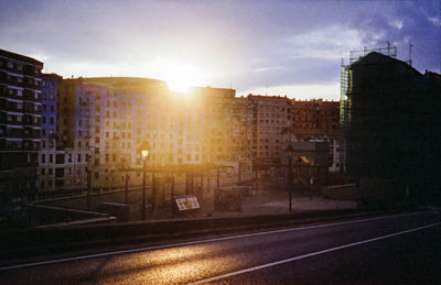 Road by buildings in city against sky during sunset