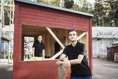 Portrait of confident carpentry student leaning on plank with classmate in background