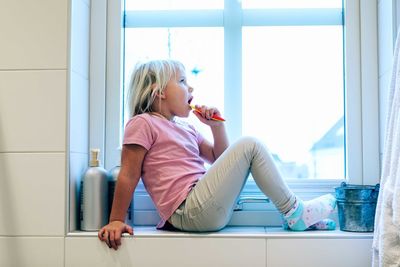 Girl brushing teeth while sitting on bathroom window