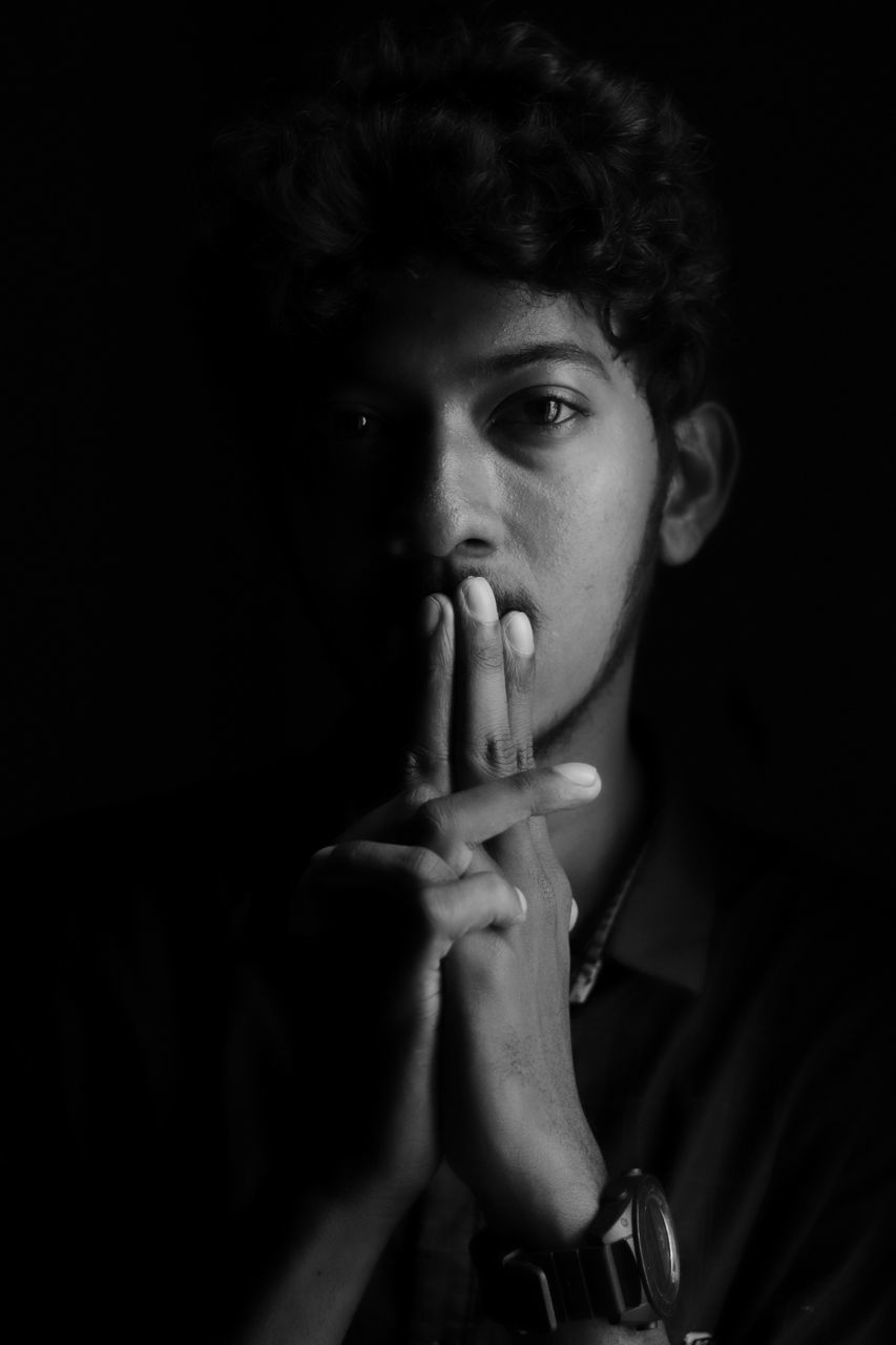 one person, portrait, young adult, looking at camera, indoors, front view, young men, social issues, headshot, real people, lifestyles, bad habit, holding, leisure activity, black background, studio shot, sign, dark, finger on lips