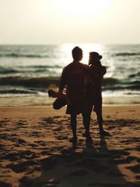 Rear view of couple standing on beach against sky during sunset