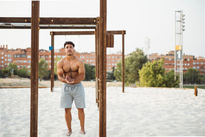 Muscular hispanic ethnic male athlete with naked torso rubbing sand on hands during training on the beach looking away