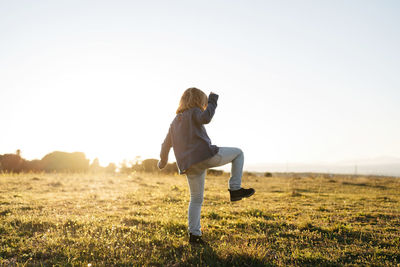 Back view of anonymous adorable active little girl in casual wear playing and dancing in green field while enjoying sunny summer evening in countryside