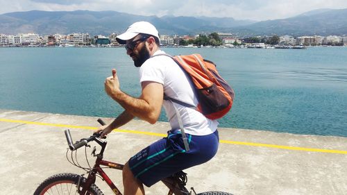 Man showing thumbs up sign while riding bicycle on road by lake in town