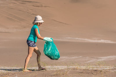 Mature woman in a white hat and green gloves collecting garbage in a green bag. concept plogging