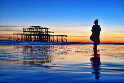 Silhouette woman standing on shore while looking at ruined west pier during sunset