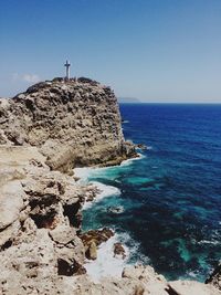 Cross on rock formation by sea against clear sky