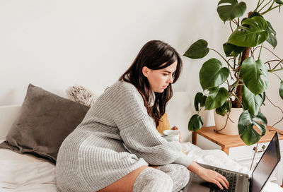 Woman in cozy sweater and warm socks sitting on bed, using laptop.