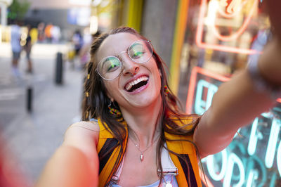 High angle of young woman with backpack and glasses looking at camera while taking selfie on modern city street