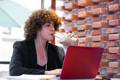 A non-binary person drinks a cold beverage while is working at the laptop