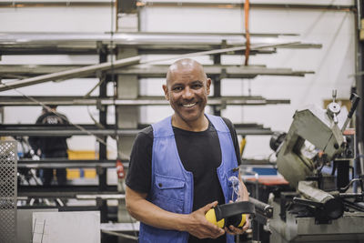 Portrait of happy bald carpenter holding ear protectors in warehouse