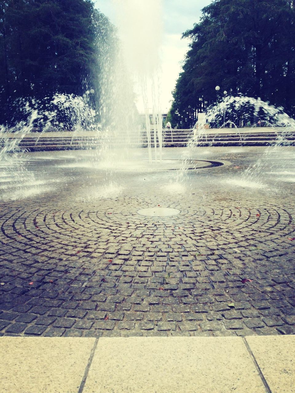 fountain, motion, built structure, building exterior, street, sunlight, day, spraying, tree, outdoors, splashing, architecture, cobblestone, water, no people, park - man made space, road, incidental people, footpath, steps