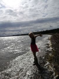 Girl with arms outstretched standing at beach against cloudy sky