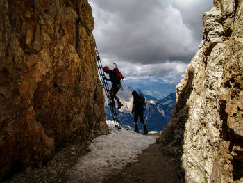 People climbing rocky mountain with ladder against cloudy sky