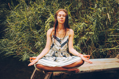 Woman practicing yoga on pier by river. young female wearing dress in lotus pose. 