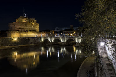 Illuminated bridge over river by buildings against sky at night