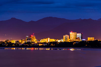 Illuminated city by sea against sky at dusk