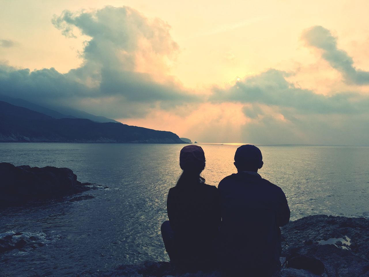 REAR VIEW OF SILHOUETTE COUPLE SITTING ON SHORE AGAINST SKY