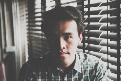 Close-up portrait of young man by window blinds at home