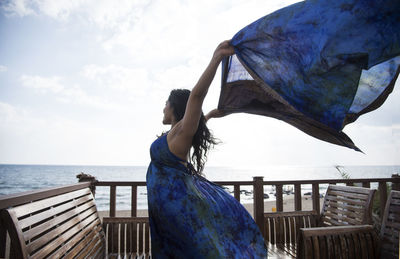 Happy woman waving dupatta while standing by wooden chairs at beach resort