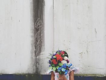 Woman covering face with bouquet against wall