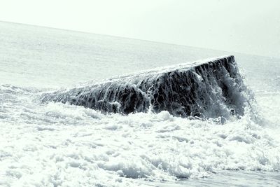 Tilt shot of wave splashing over rock on shore against sky