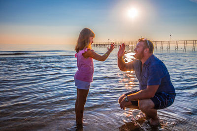 Girl giving high five to father while standing in sea against sky during sunset