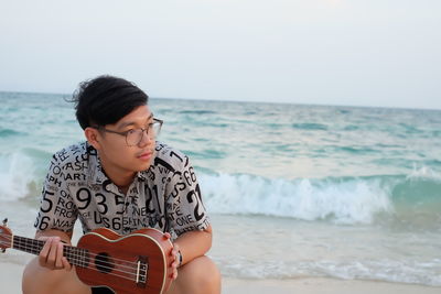 Young man holding violin while sitting at beach against sky during sunset