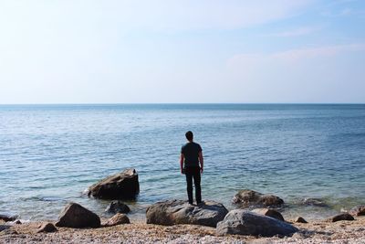 Rear view of man sitting on rock by sea against clear sky
