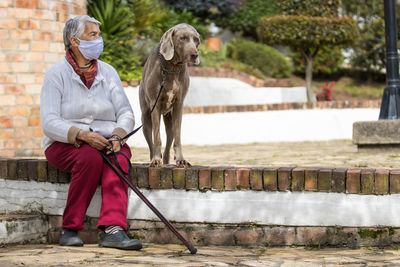 Full length of senior wearing mask with dog sitting outdoors