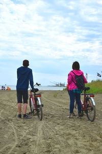 Rear view of friends with bicycle standing at beach against sky