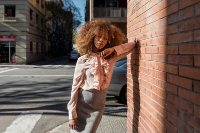 Portrait of beautiful young woman with afro hairdo leaning against brick wall in the city