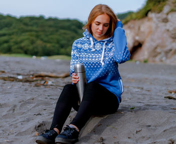 Young woman holding insulated drink container while sitting at beach