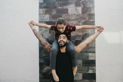 Daughter with arms outstretched sitting on father's shoulder against wall