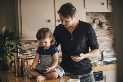 Daughter using smart phone while father standing in domestic kitchen