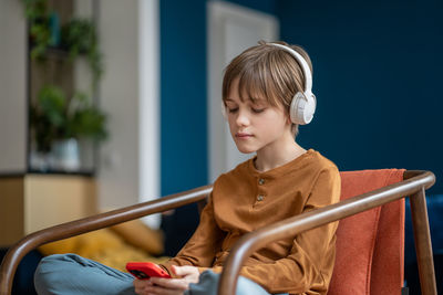 Portrait of music lover teen boy listening to music in headphones sitting on armchair at home.