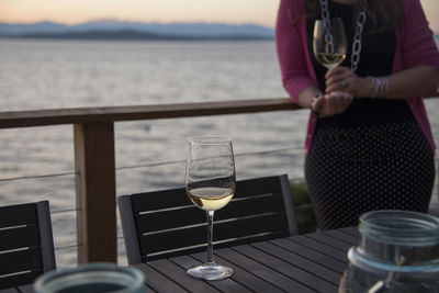 Midsection of woman holding wineglass at restaurant by sea during sunset