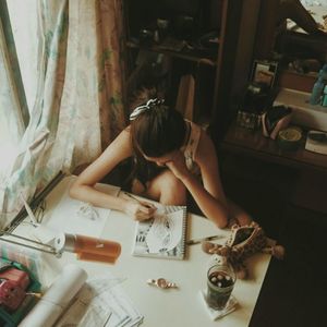 High angle view of young woman drawing on book at table by window in home