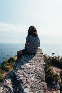 Rear view of man sitting on rock by sea against sky