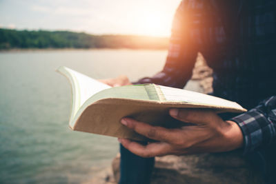 Midsection of person reading book against lake