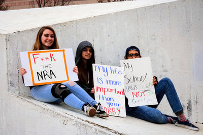 Full length portrait of friends with placard messages sitting on retaining wall