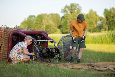 Boy assisting pilot in preparing hot air balloon on grassy field during sunset