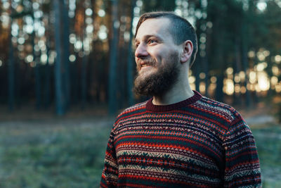 Young man looking away while standing against trees in forest